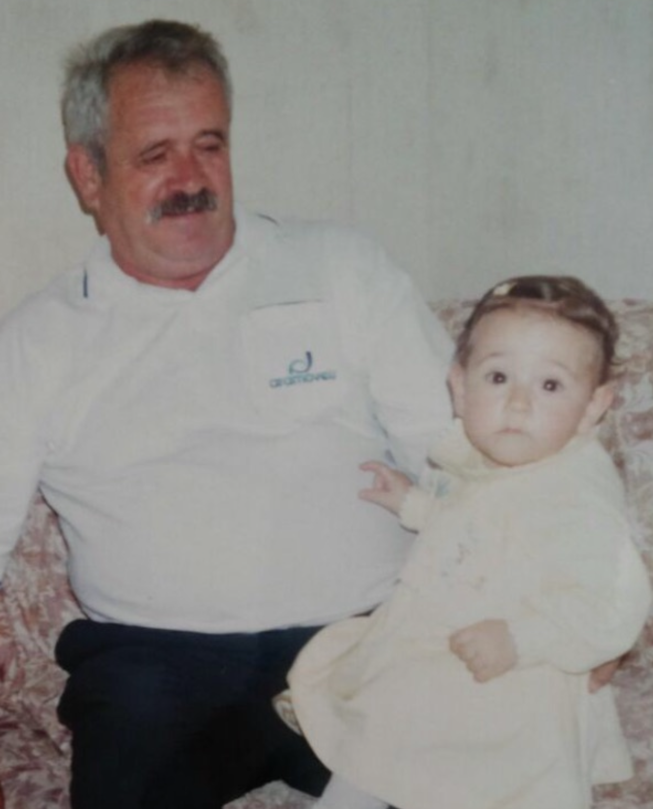 Vintage photo of a grandfather lovingly holding his granddaughter on a floral couch, the inspiration behind the name FIOCA and the warmth it represents