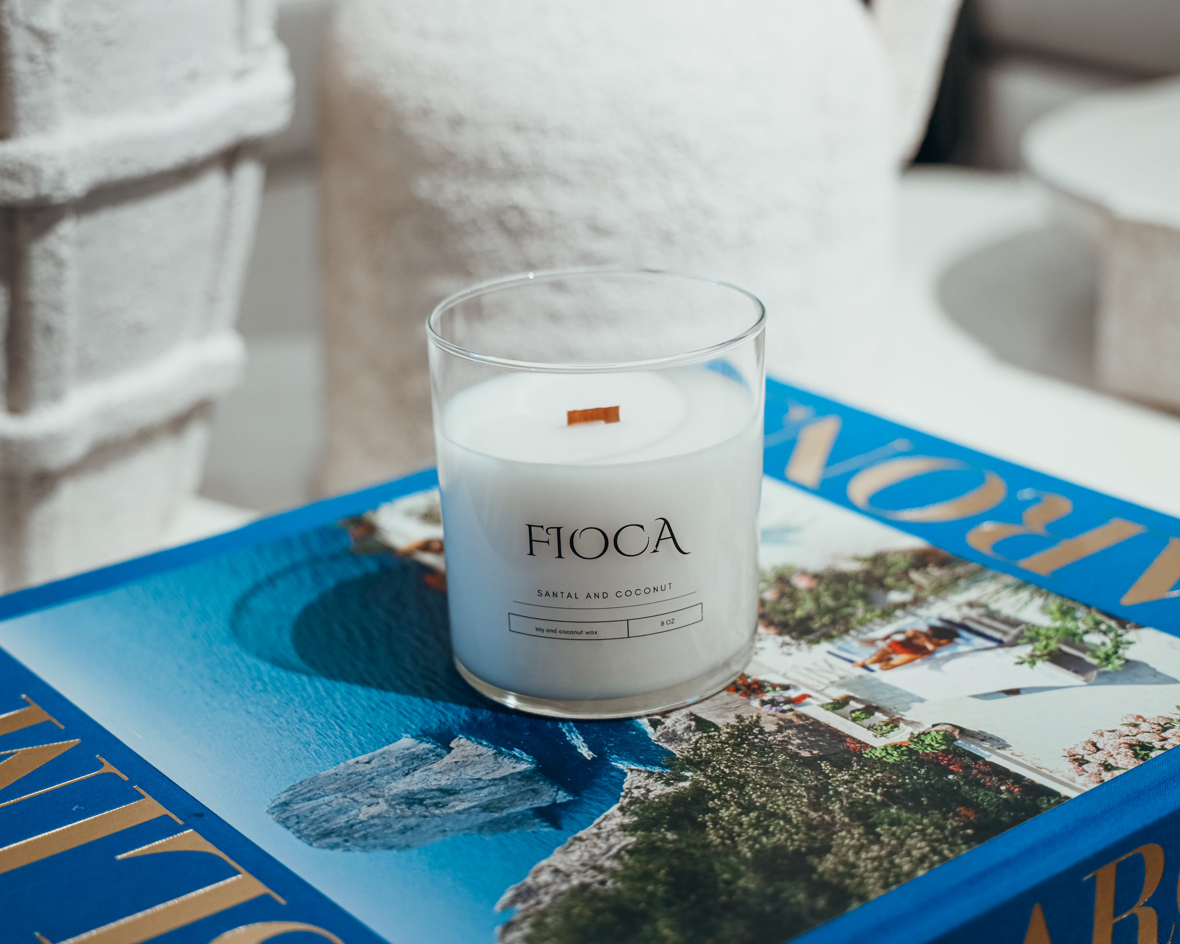 Fioca Santal and Coconut candle resting on a vibrant travel book, blending luxury fragrance with bright, coastal aesthetics